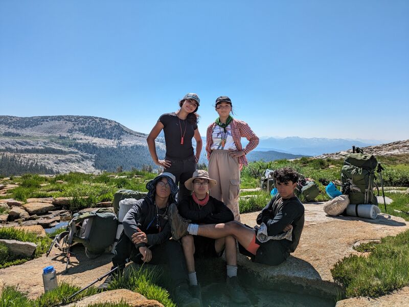 A group of nine young adults stands on a rocky, mountainous terrain under a clear blue sky. Some are wearing backpacks, suggesting they are hiking or camping. The landscape features sparse vegetation and distant mountain ranges. The group appears to be diverse and is posing for a photo, with some making peace signs. The overall impression is one of adventure and camaraderie in a natural setting.
